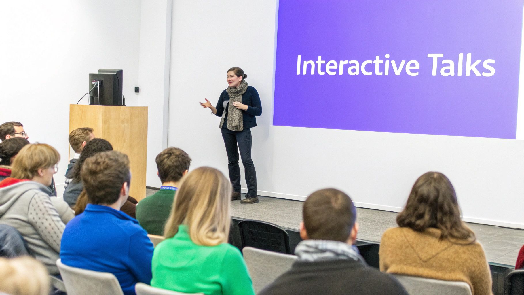 A woman presents 'Interactive Talks' on a screen to an attentive audience in a lecture hall.