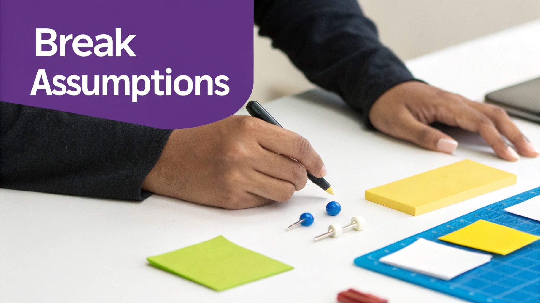 Hands holding a pen over a desk with sticky notes and creative tools, promoting 'Break Assumptions'.