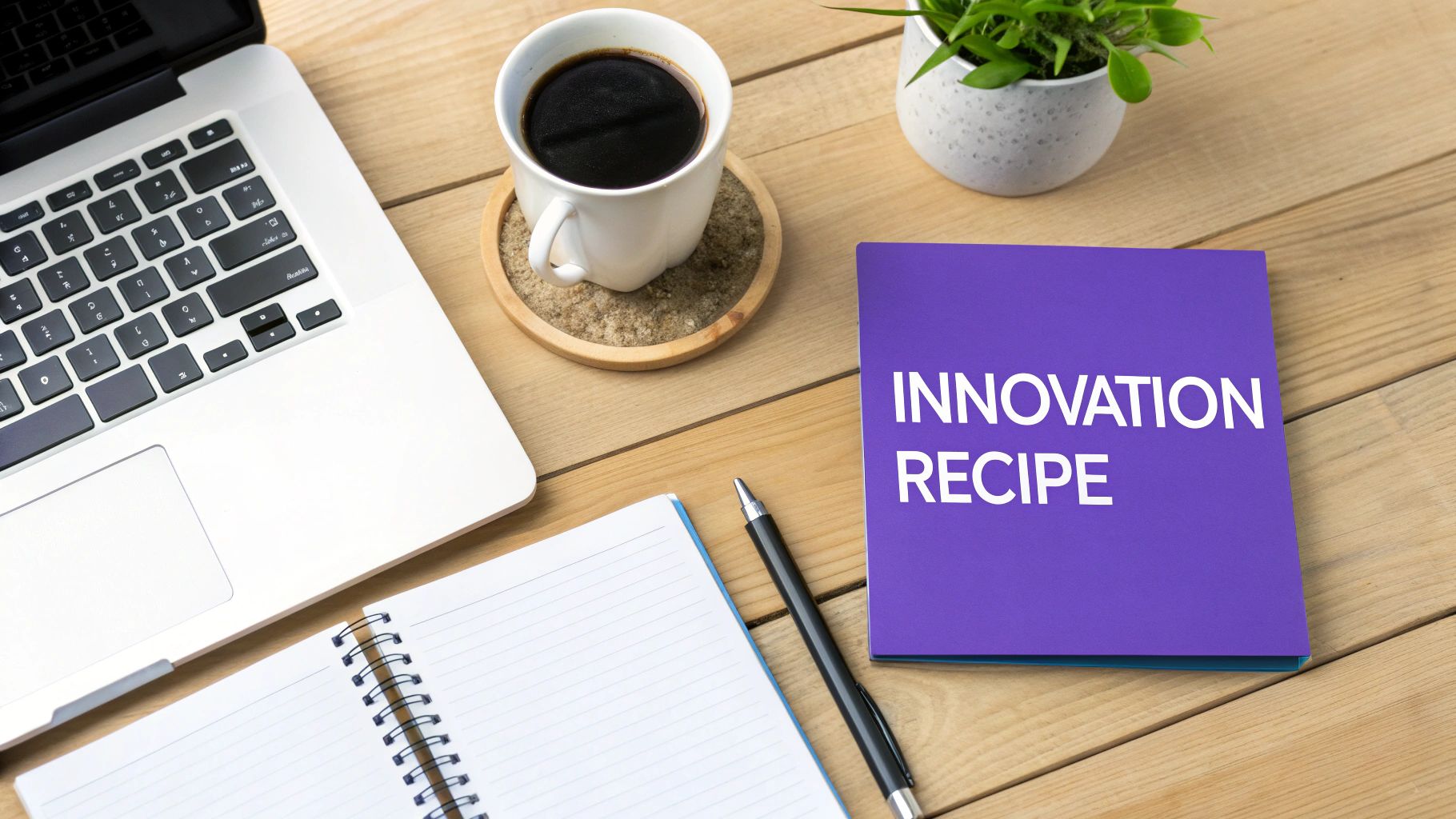A desk flat lay with a laptop, coffee, plant, notebook, pen, and an 'INNOVATION RECIPE' folder.