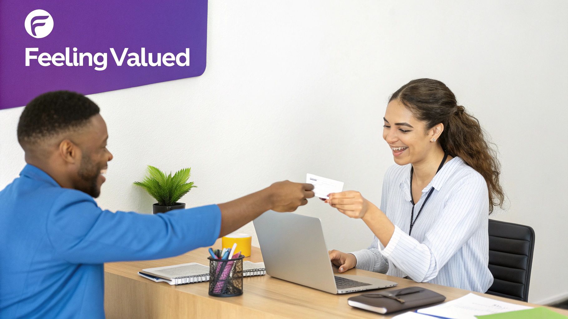 Two smiling colleagues exchanging a white card at a desk in an office setting.