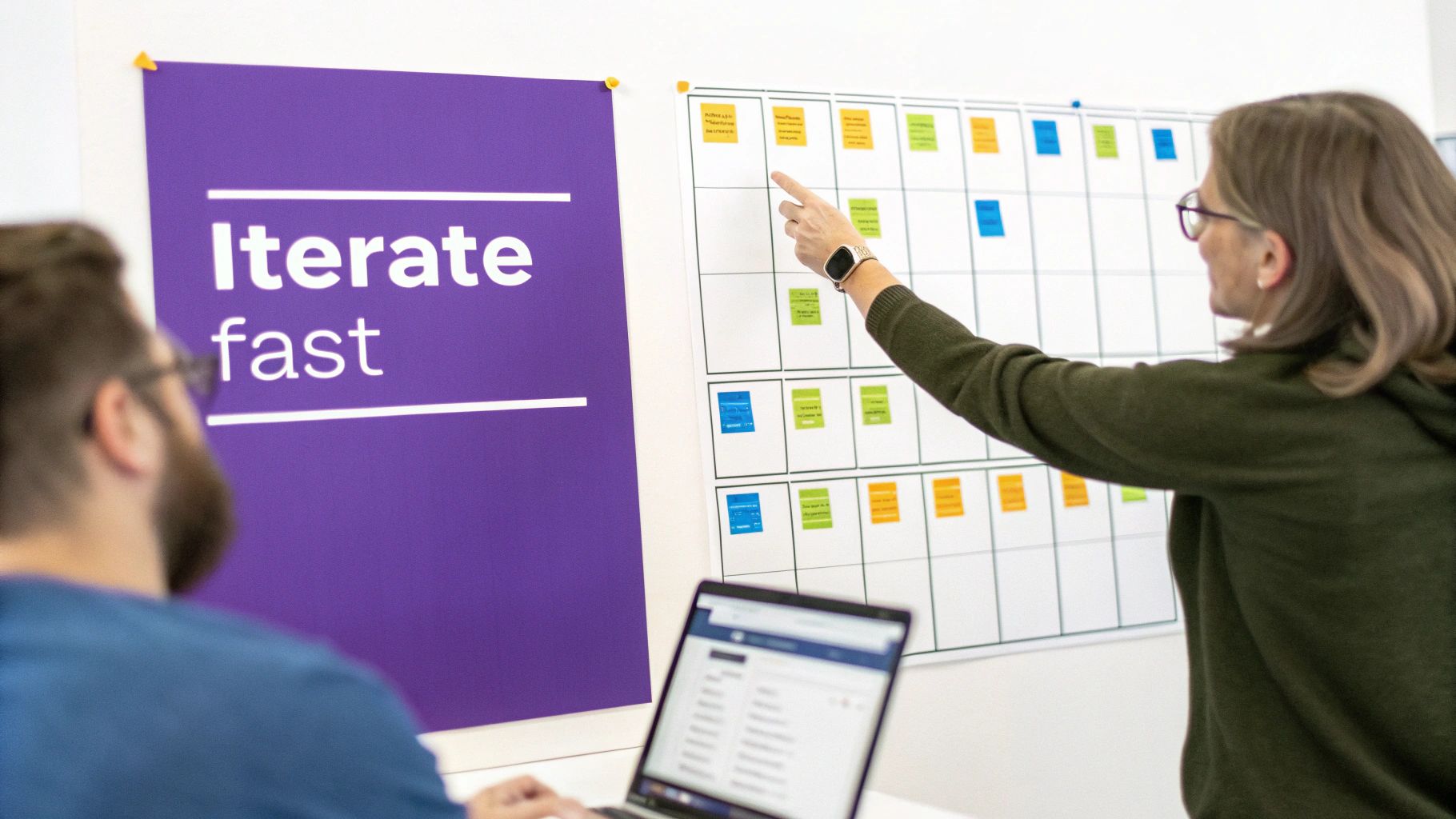 Two colleagues in an office collaborate, one pointing at a whiteboard with sticky notes and an 'Iterate fast' poster.