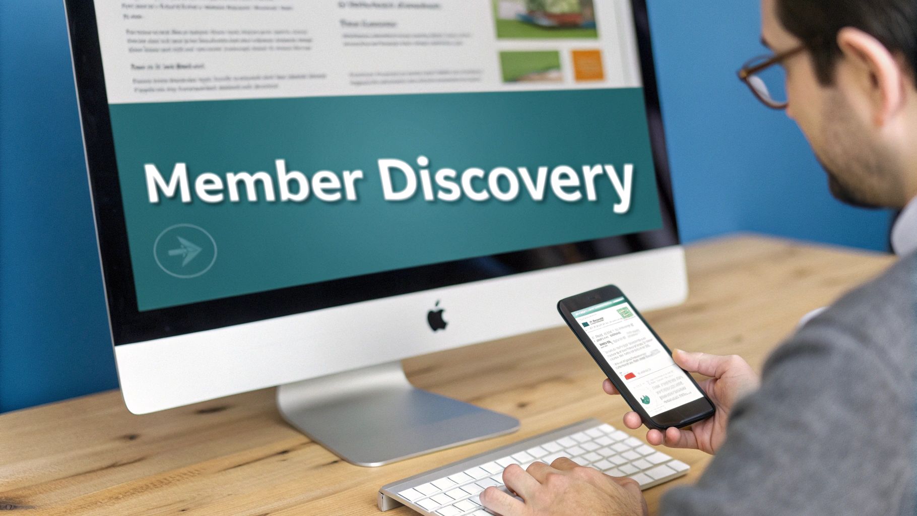 A person views "Member Discovery" on an iMac while holding a smartphone and typing on a keyboard.