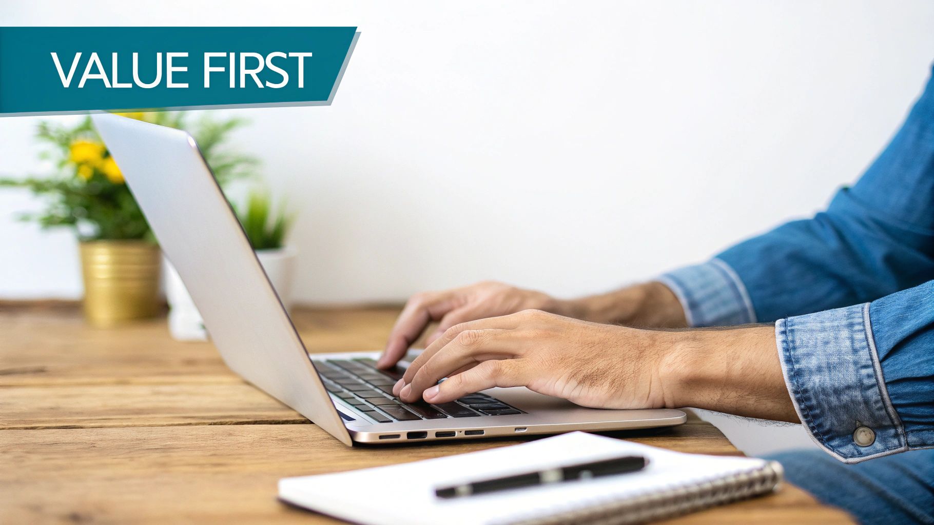 A person in a denim shirt types on a laptop, with 'VALUE FIRST' banner, on a wooden desk with plants.