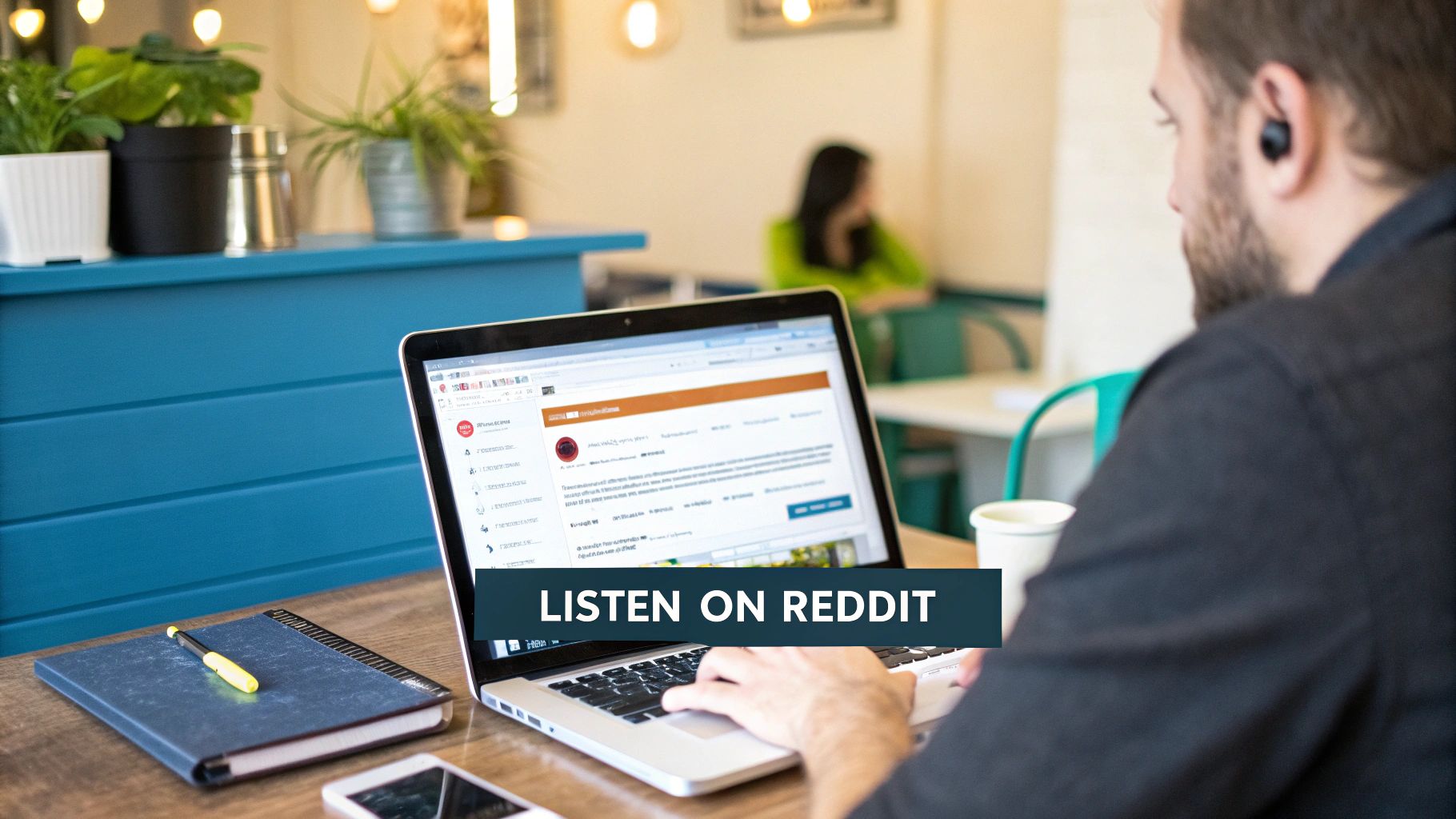 Man with an earbud working on a laptop displaying Reddit, with a notebook and phone on the desk.