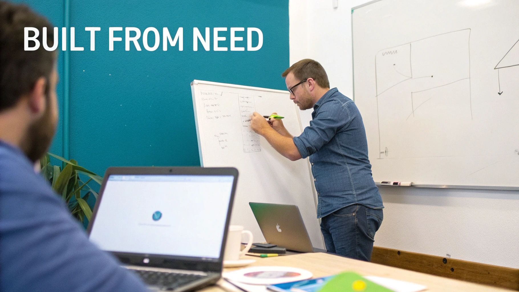 A man in glasses writes on a whiteboard while another person works on a laptop in an office.