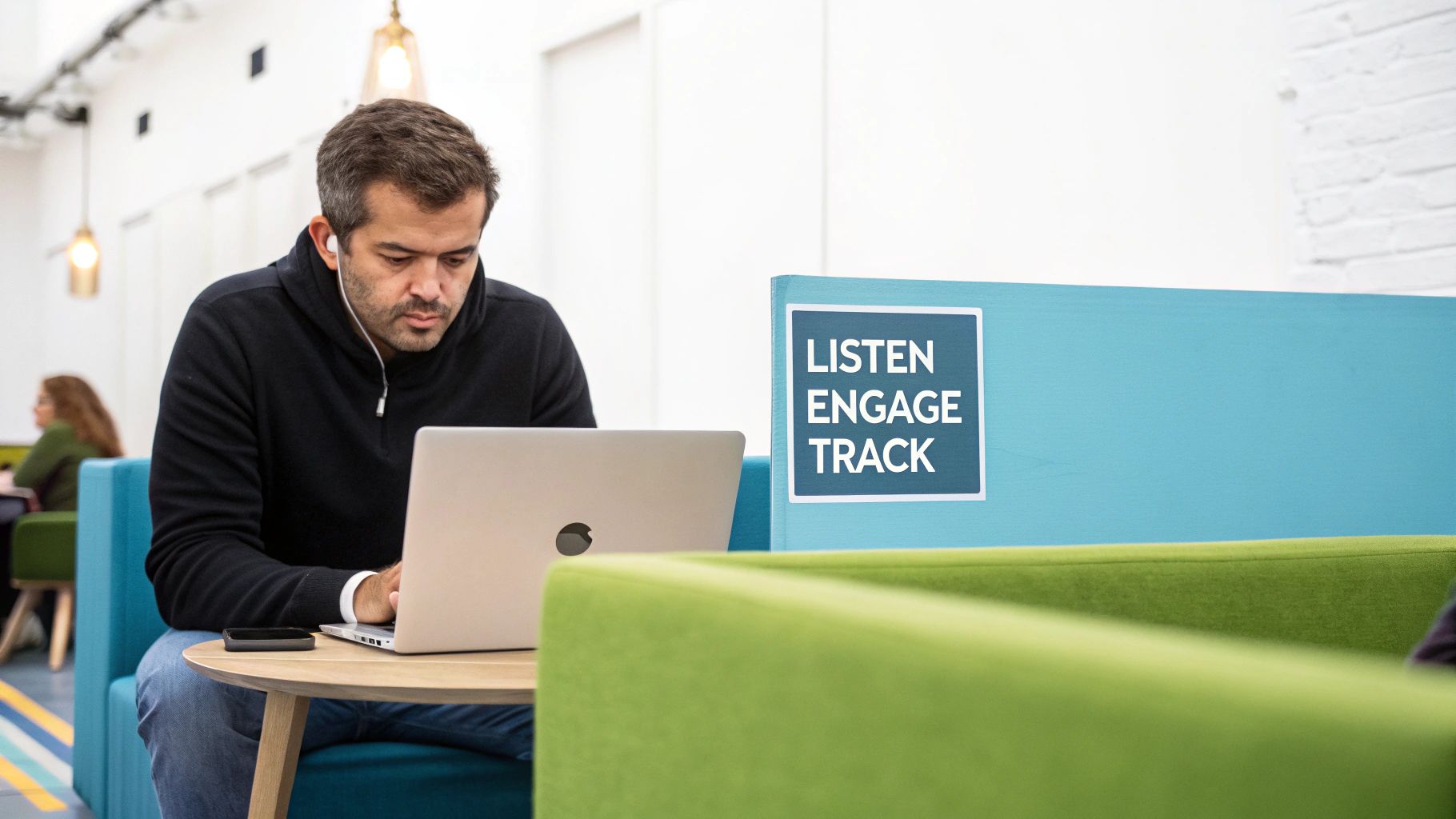 A man with earbuds working on a laptop in a bright office lounge, with a sign 'LISTEN ENGAGE TRACK'.