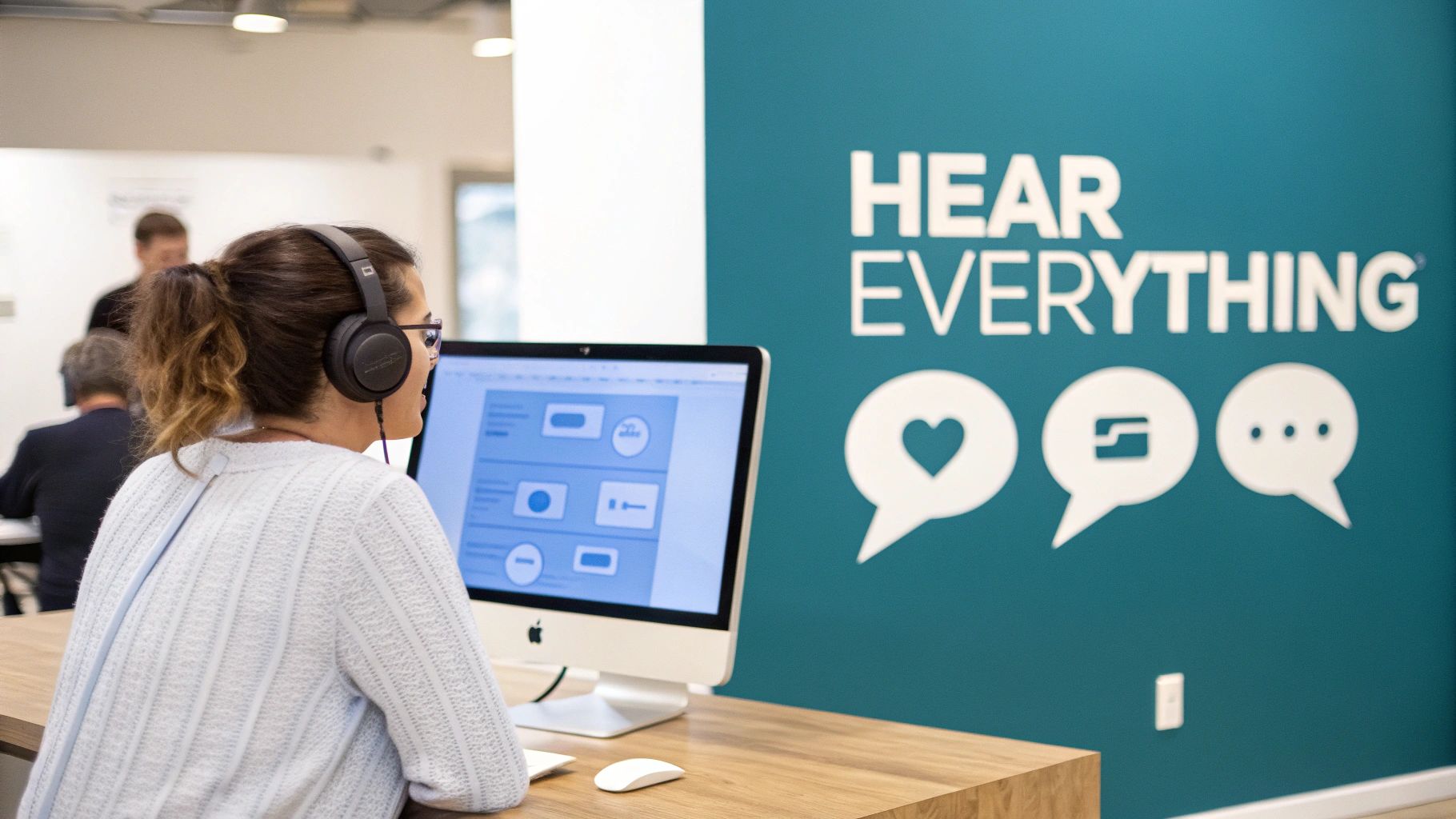 A woman with headphones works on an iMac in front of a wall promoting 'Hear Everything'.