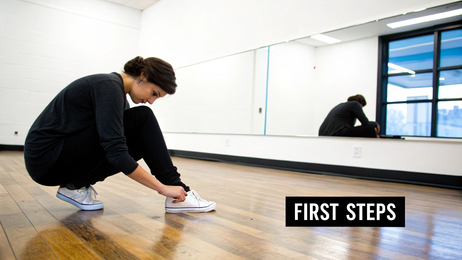 A woman in a dance studio squats down, tying her white shoe, with her reflection in a mirror.