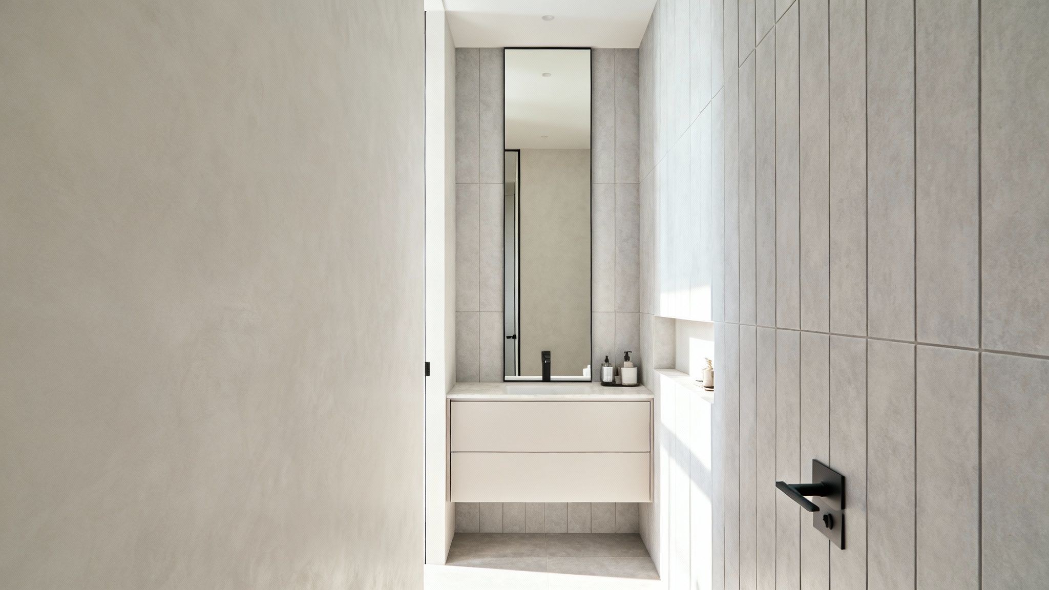 Modern, narrow bathroom with light grey tiles, a black-framed mirror, and a sleek vanity.