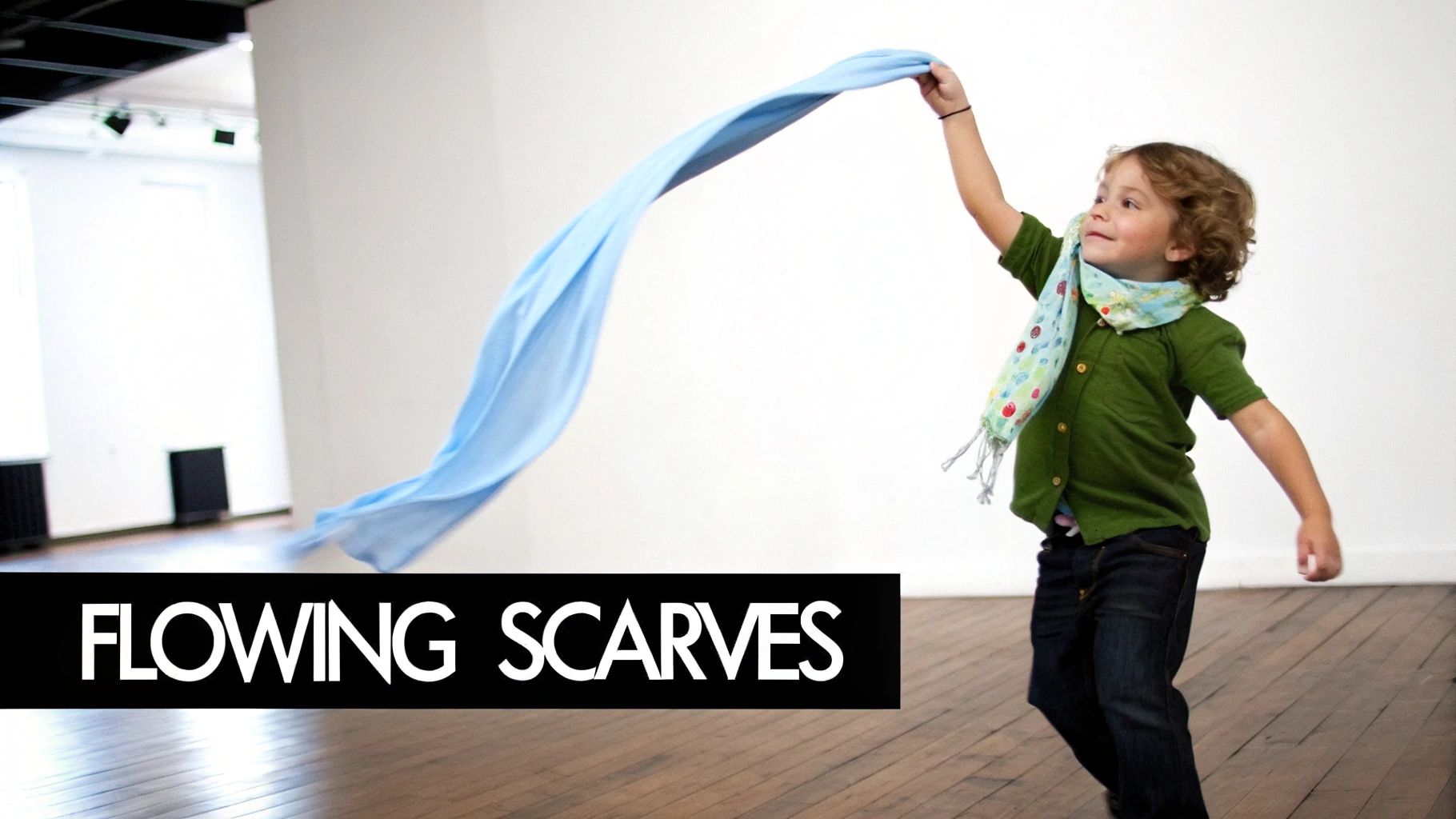 A joyful child in a green shirt and patterned scarf plays with a flowing light blue scarf indoors.