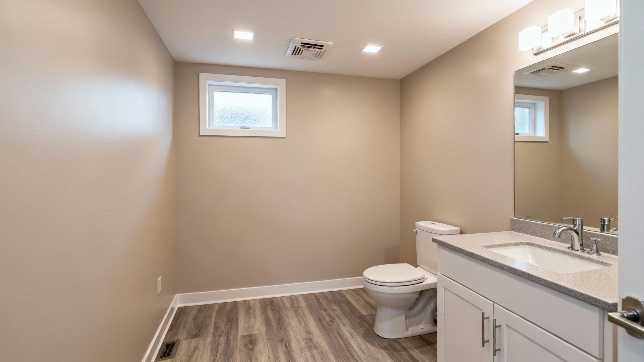 A clean and modern basement bathroom featuring a toilet, vanity, mirror, and small window.
