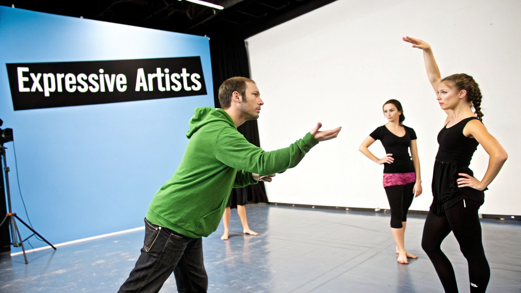 A male instructor in a green hoodie gestures towards three female dancers in a studio with an 'Expressive Artists' banner.