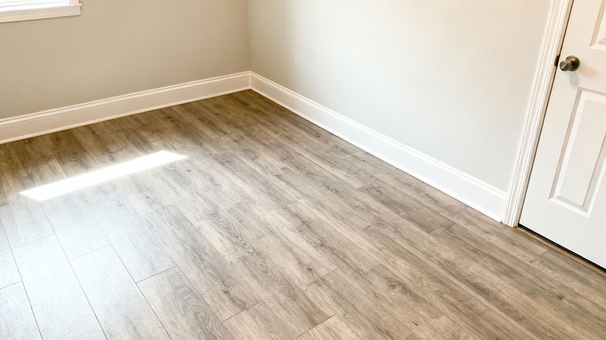 An empty room showcasing light wood-look flooring, light grey walls, white trim, and a door, with sunlight.
