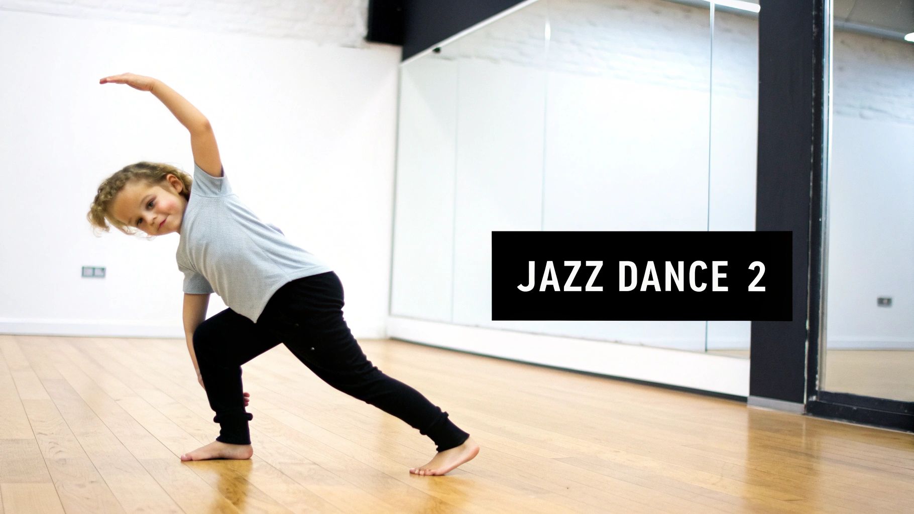 A young child with curly hair performs a jazz dance pose in a studio with mirrors.