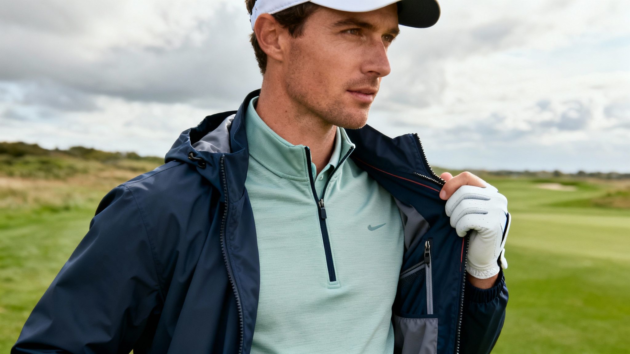 A male golfer in a navy jacket and mint green quarter-zip shirt on a golf course.