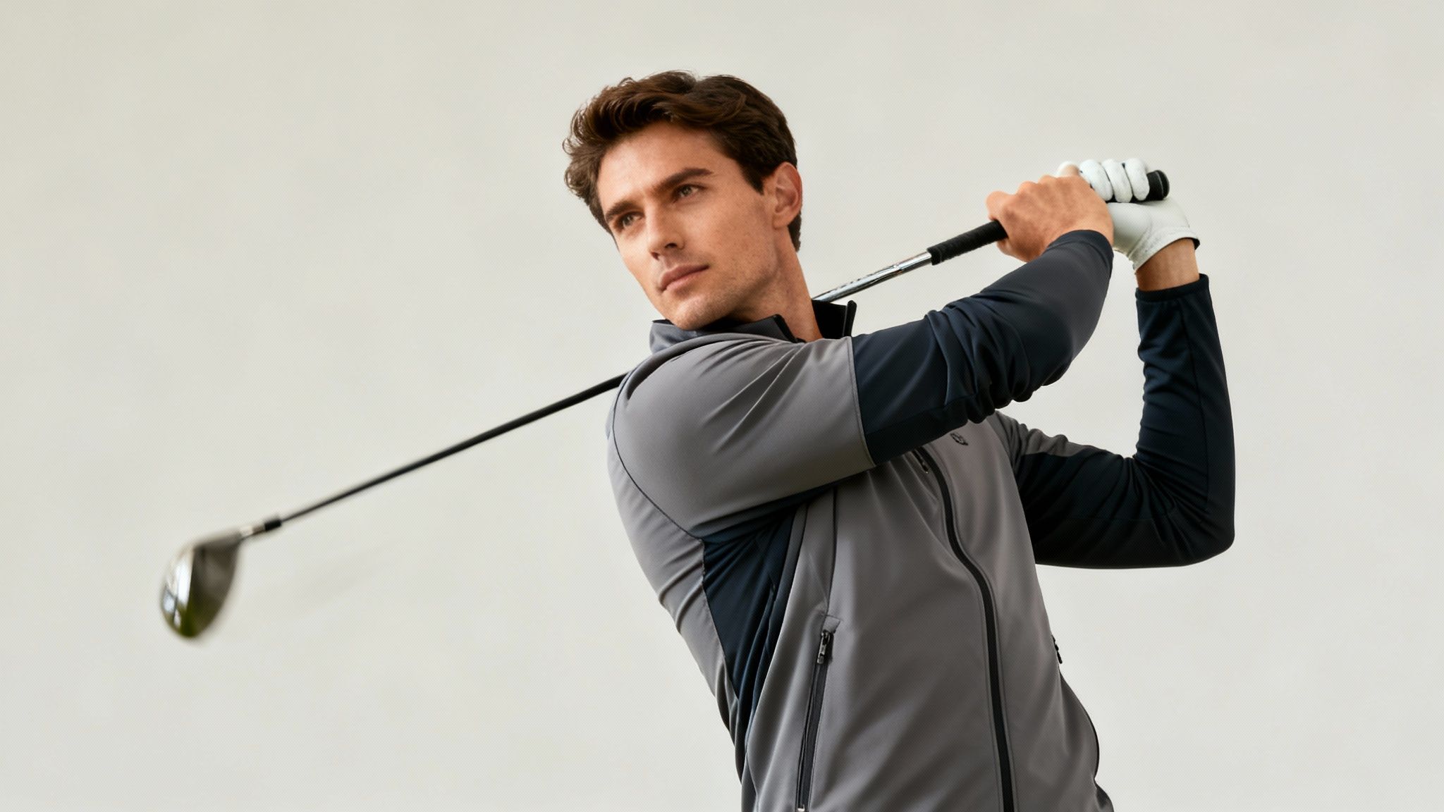 A man in a stylish grey and navy golf windbreaker mid-swing with a golf club.