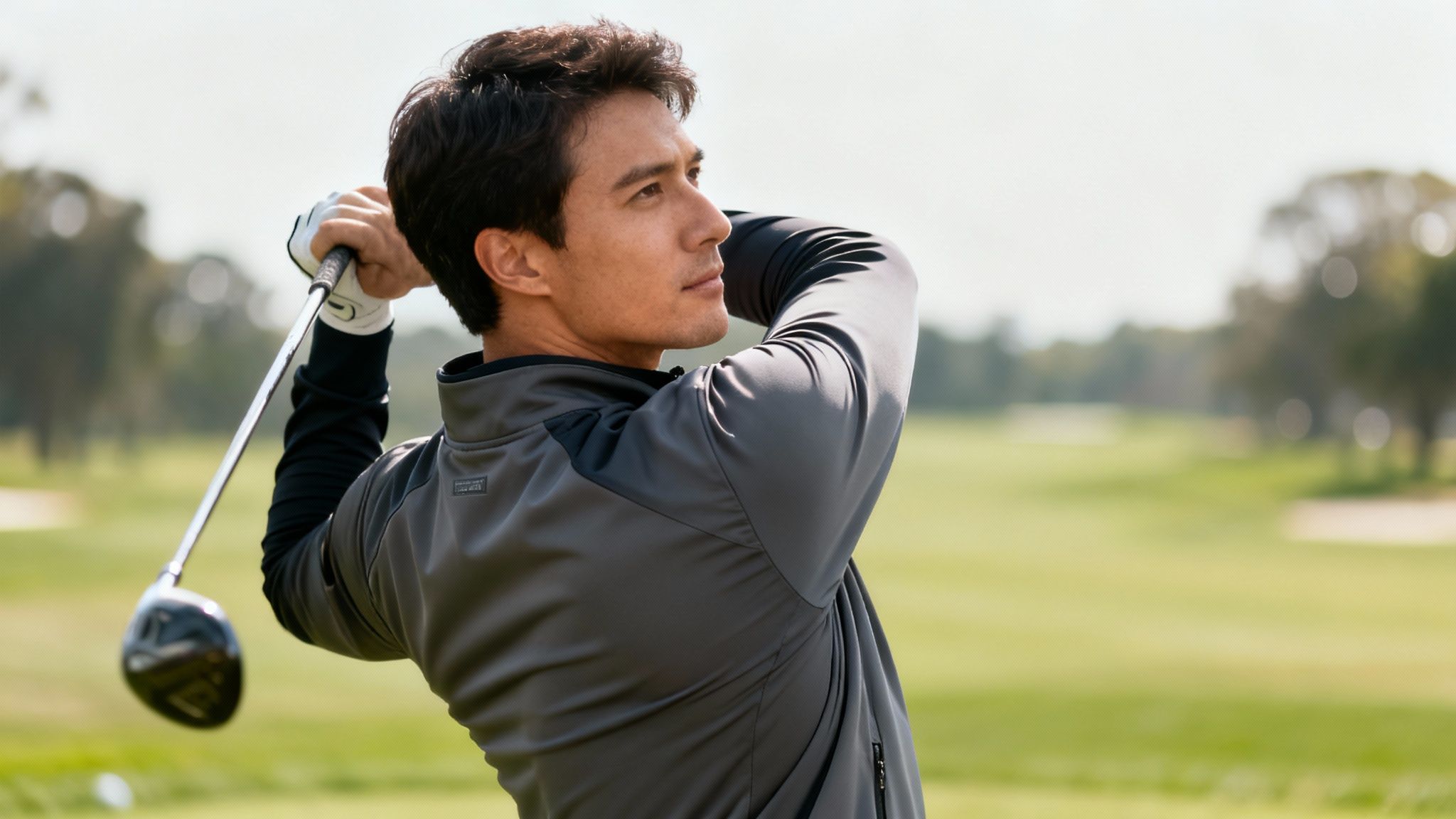 A man in a grey golf windbreaker jacket swings a golf club on a green course.