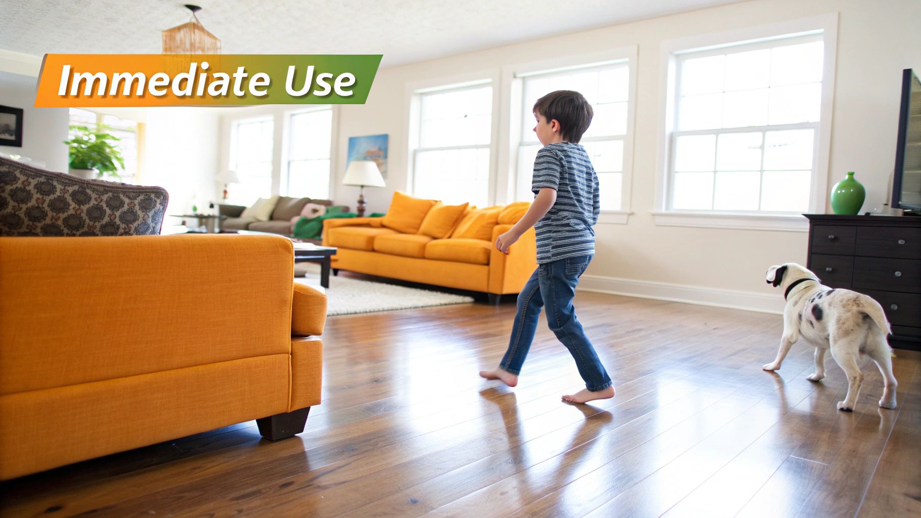 A young boy walks barefoot on a shiny wooden floor, followed by a dog in a bright living room.