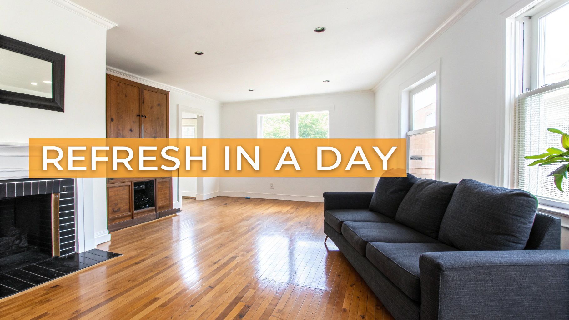 A bright, freshly renovated living room with shiny hardwood floors, a dark grey sofa, a fireplace, and built-in wooden cabinets.