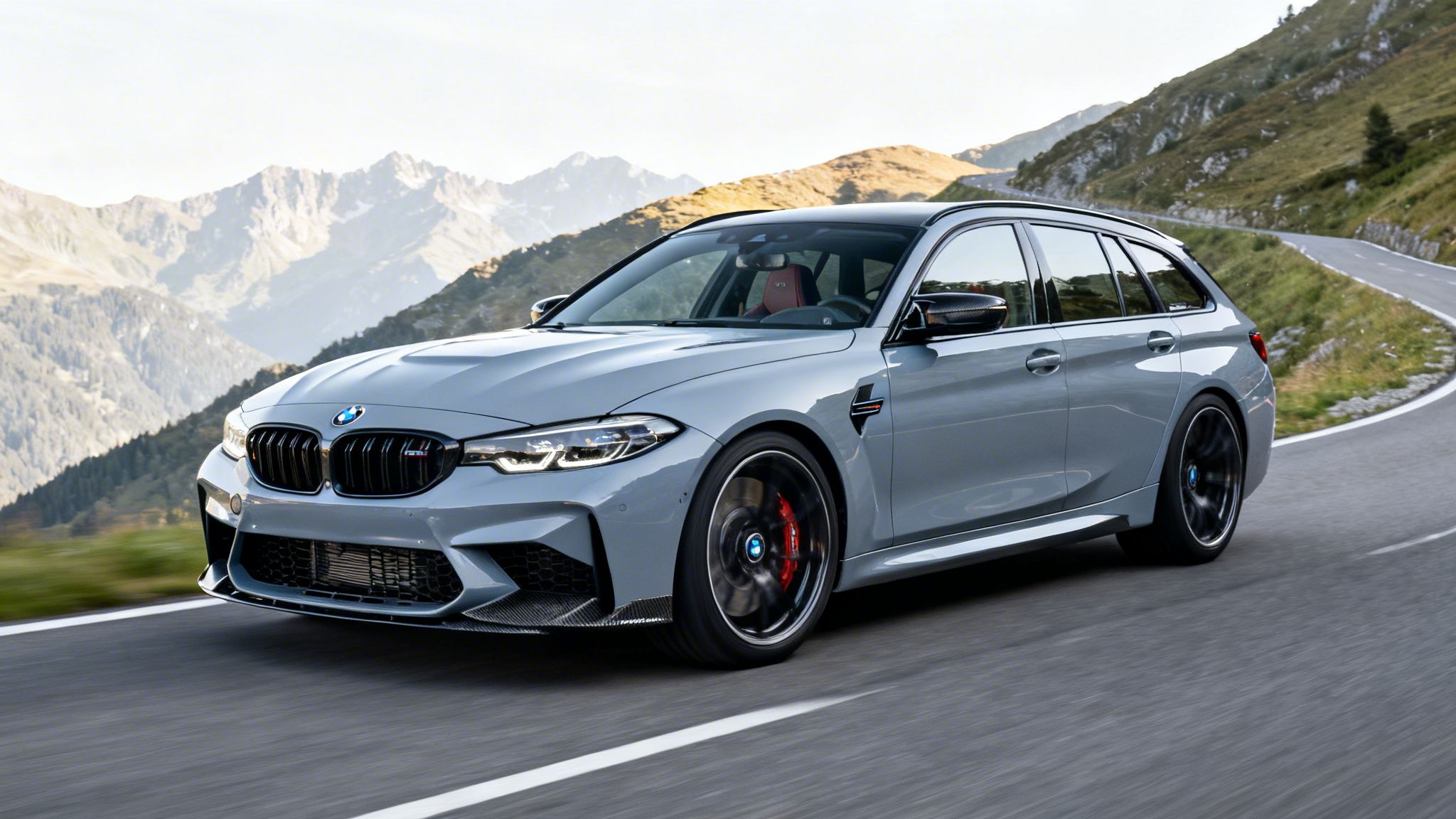 Un BMW M3 Touring Competition gris conduce por una carretera sinuosa de montaña al atardecer, con picos al fondo.