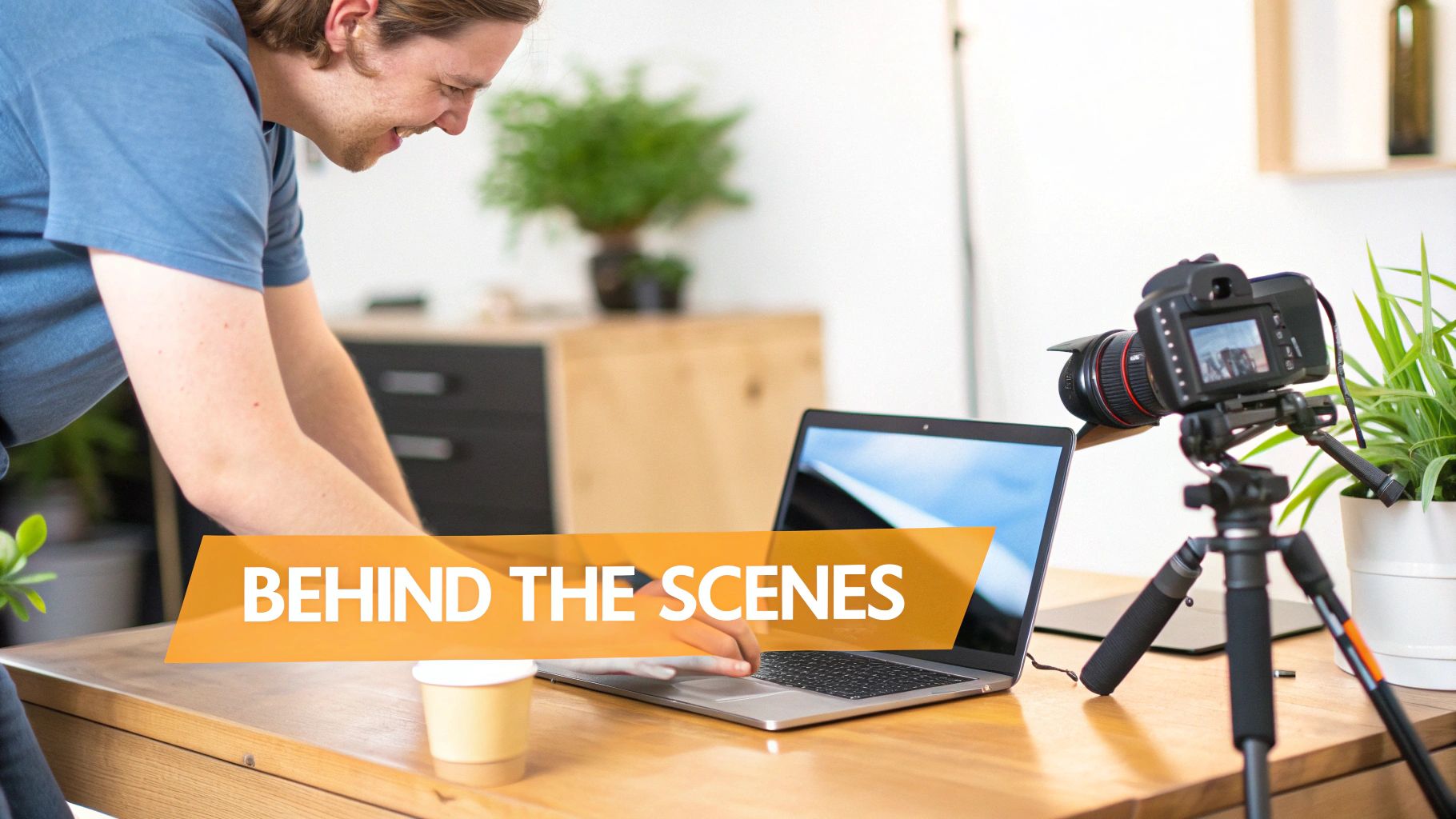 A man smiles while working on a laptop, with a camera filming, representing behind-the-scenes content creation.