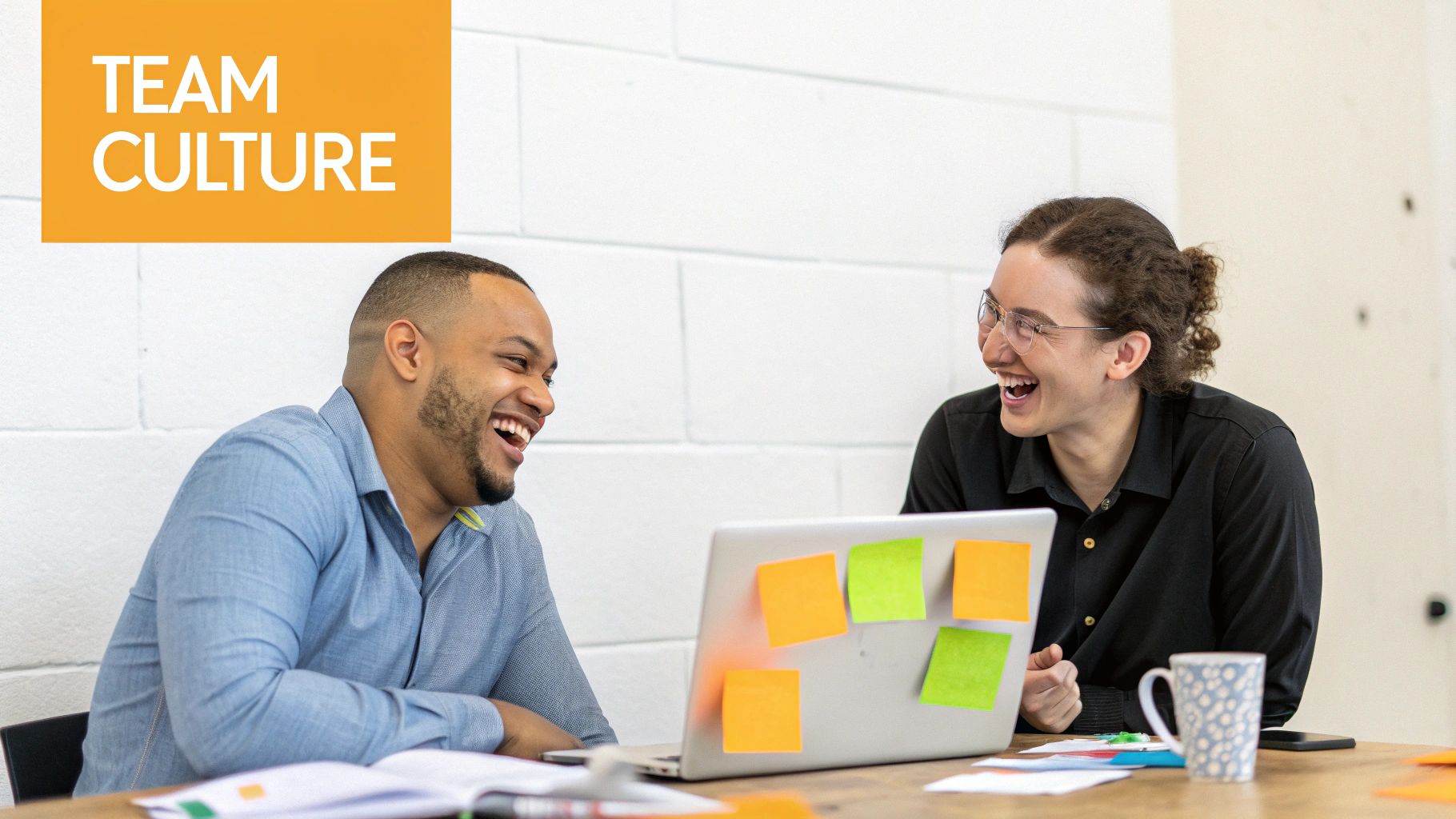 Two diverse colleagues laugh happily while collaborating at a desk with a laptop, showcasing positive team culture.