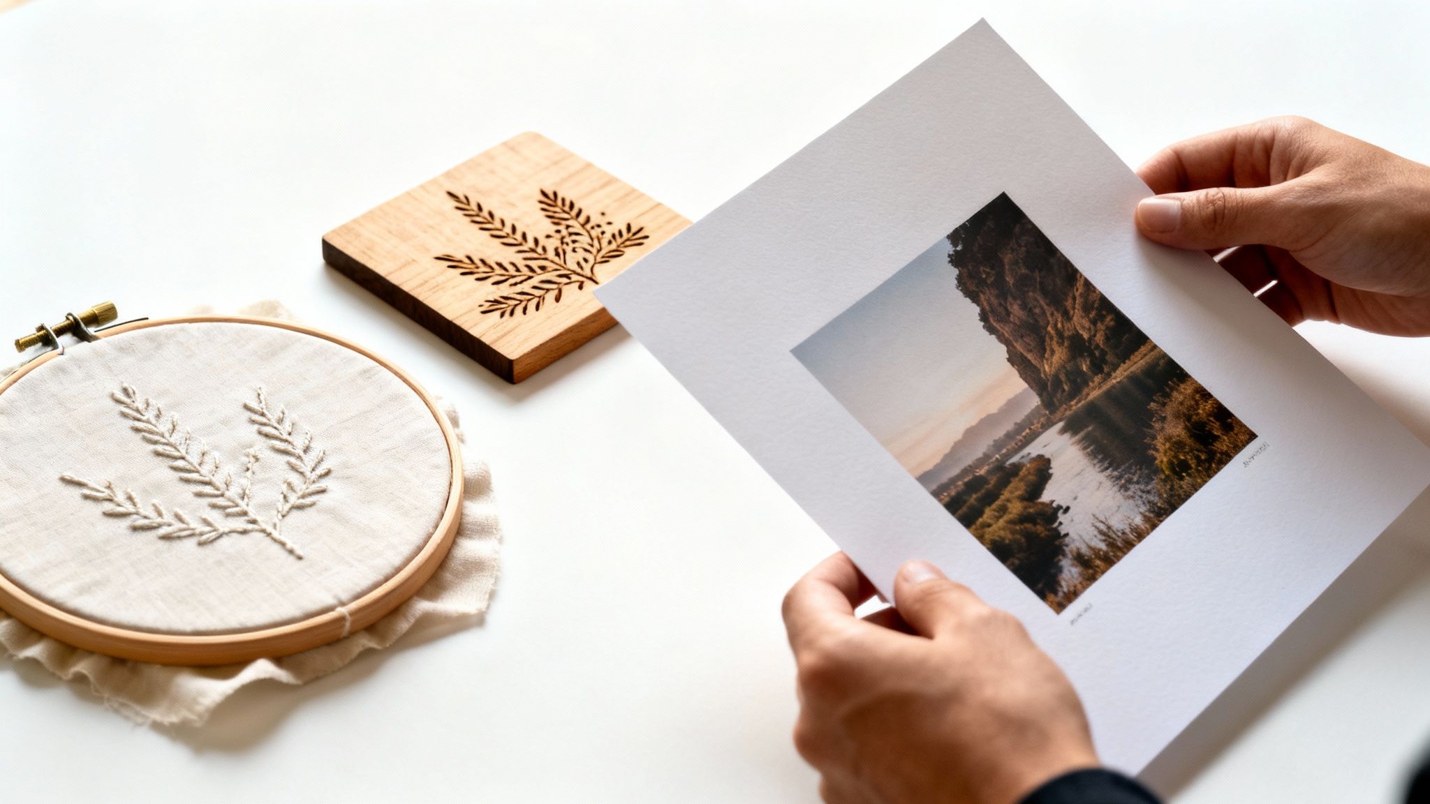 Hands holding a landscape photo, next to a wooden coaster and an embroidery hoop with plant designs.