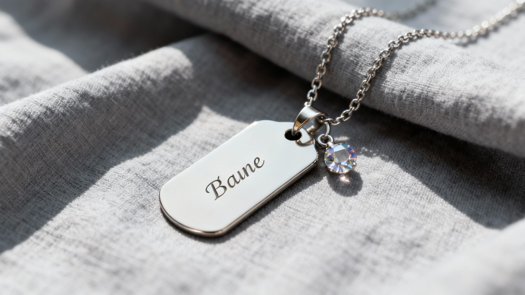 Personalized silver dog tag necklace engraved with "Baine" and a crystal charm on a grey fabric.