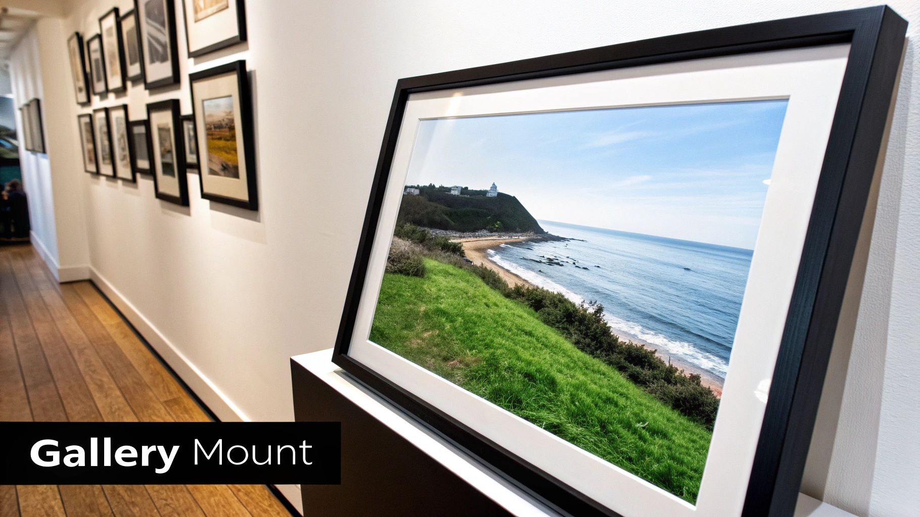 A black-framed picture of a lighthouse on a green cliff overlooking a beach and ocean in a gallery.