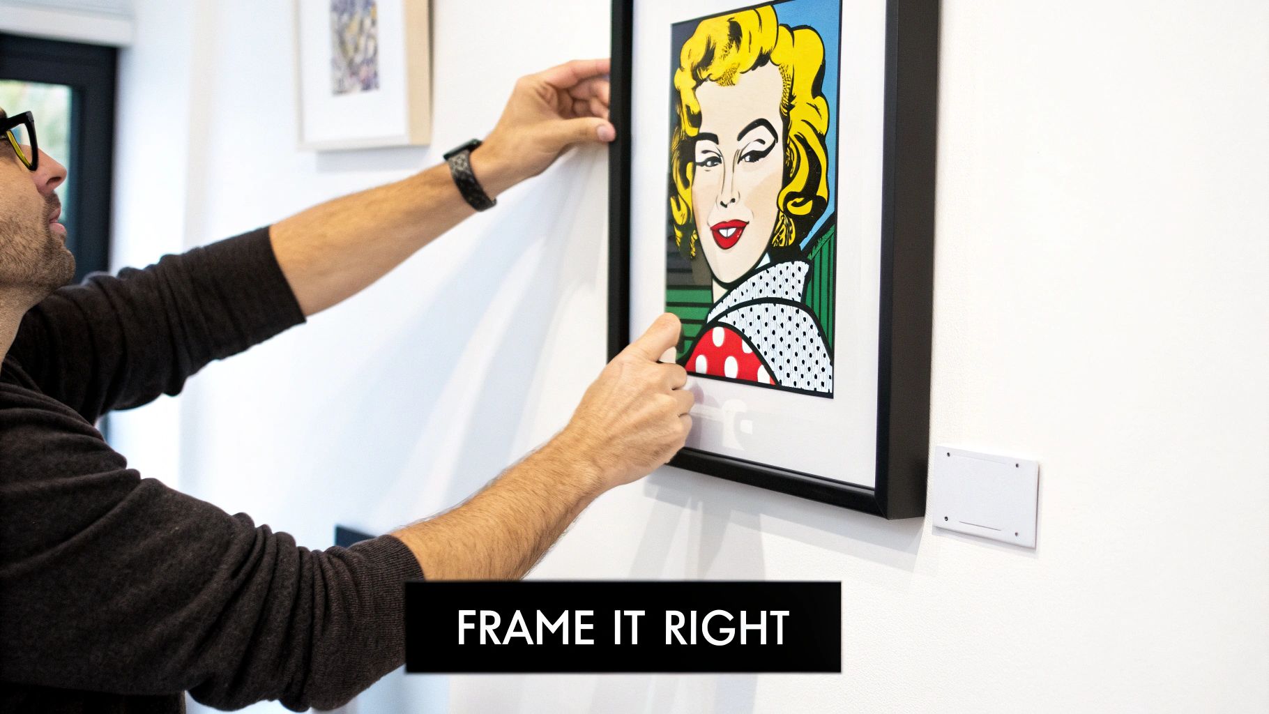A man's hands carefully hang a framed pop art print of a blonde woman on a white wall.