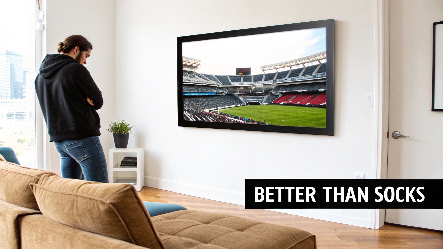 A man in a black hoodie watches a large wall-mounted TV showing a sports stadium, in a modern living room.