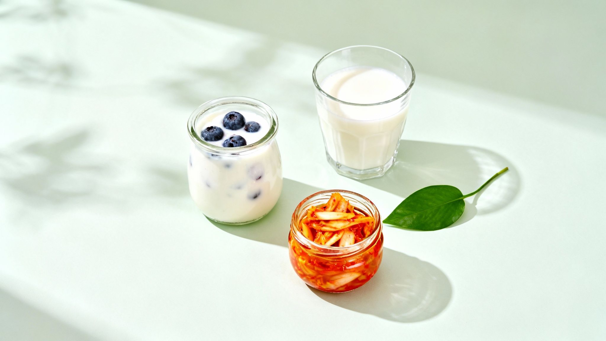 Three gut-friendly foods: blueberry yogurt, milk, and kimchi, arranged on a light green surface.