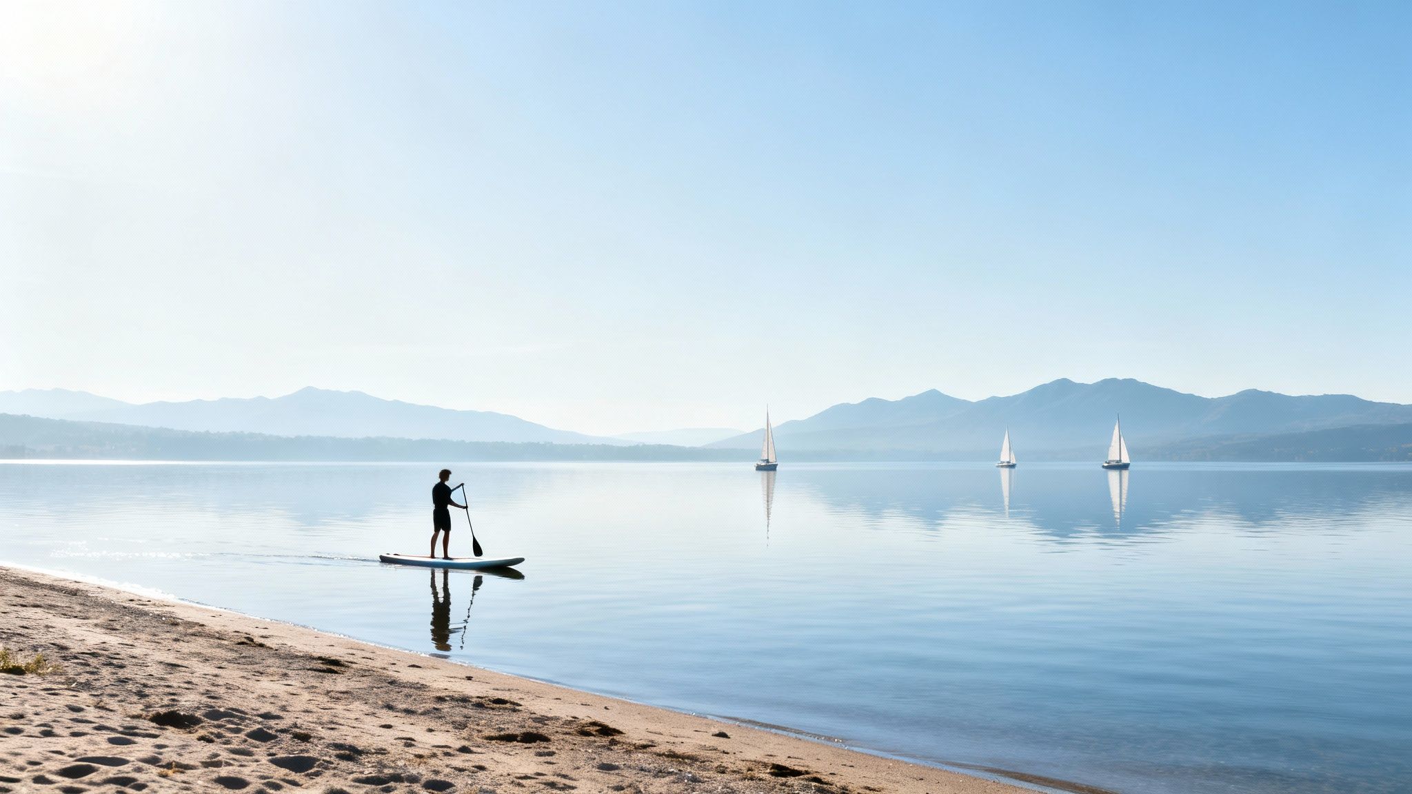 A person paddleboarding on a calm lake near a sandy beach with mountains and sailboats.