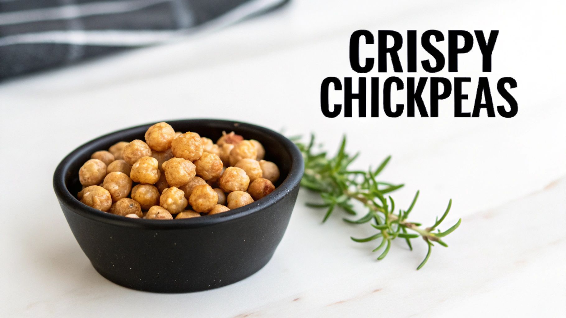 A close-up of a black bowl filled with crispy, golden-brown roasted chickpeas, garnished with fresh rosemary on a white surface.