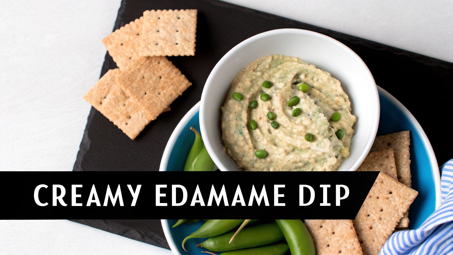 A creamy edamame dip in a white bowl, garnished with edamame beans, served with crackers and snap peas.