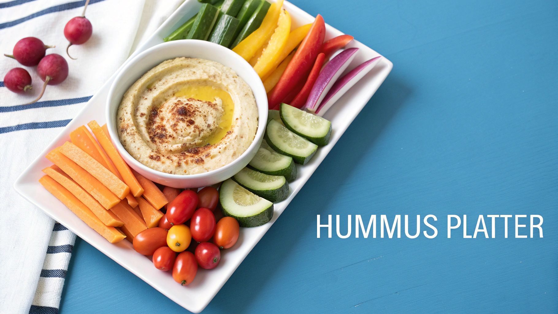A colorful hummus platter on a blue background with a bowl of hummus and assorted fresh vegetables for dipping.
