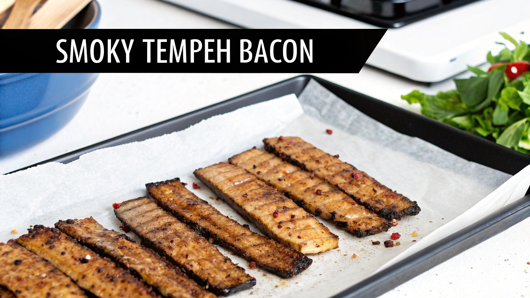 Close-up of crispy Smoky Tempeh Bacon strips on a baking sheet, ready to serve, with fresh greens in the background.
