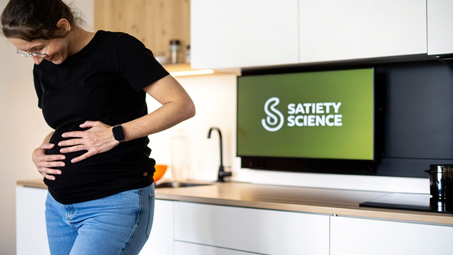 A smiling pregnant woman gently holds her belly in a modern kitchen with a 'Satiety Science' screen.