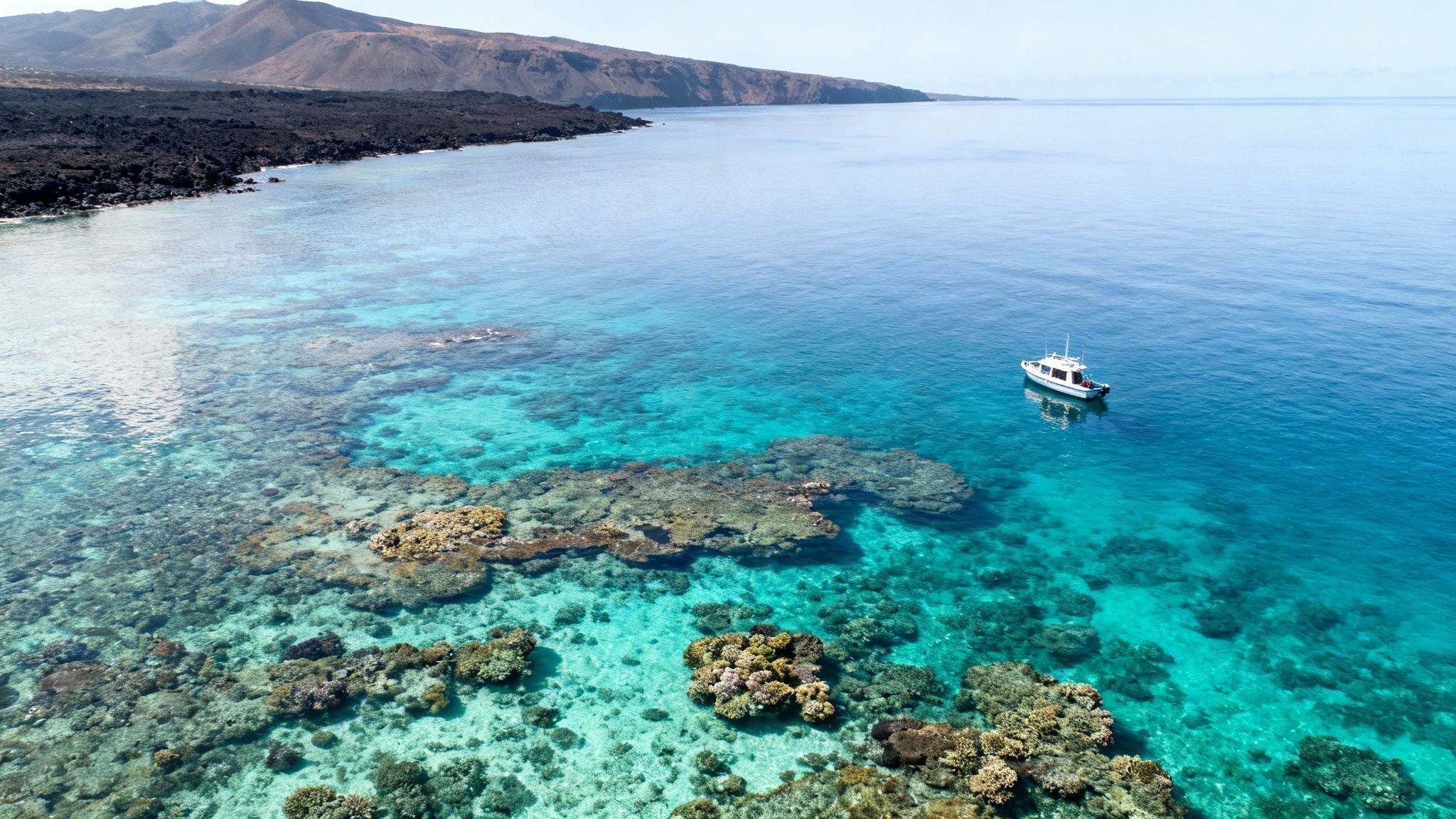 Aerial view of a white boat in clear turquoise water over a vibrant coral reef near a volcanic coastline.