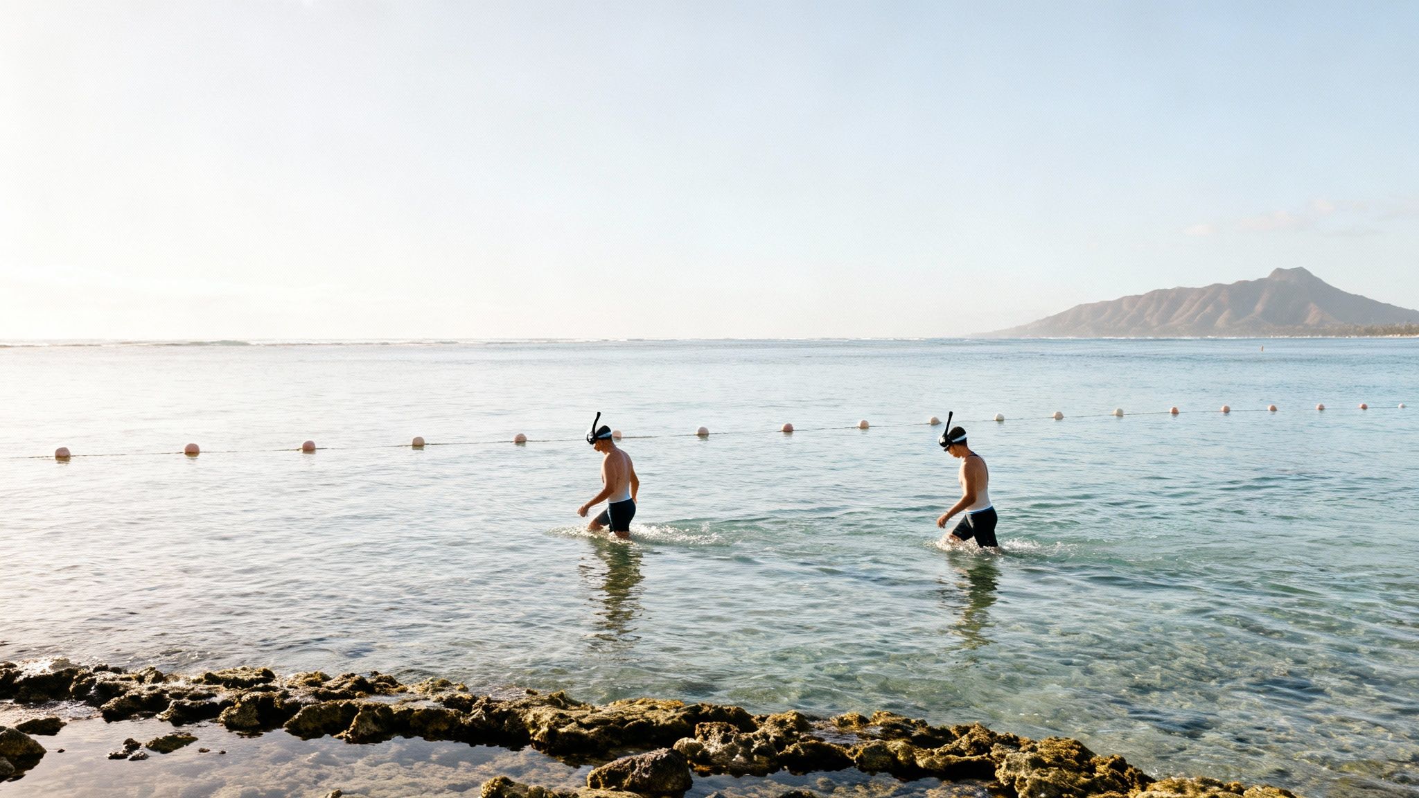 Two men with snorkels walk through clear ocean water near a rocky shore under a bright sky.