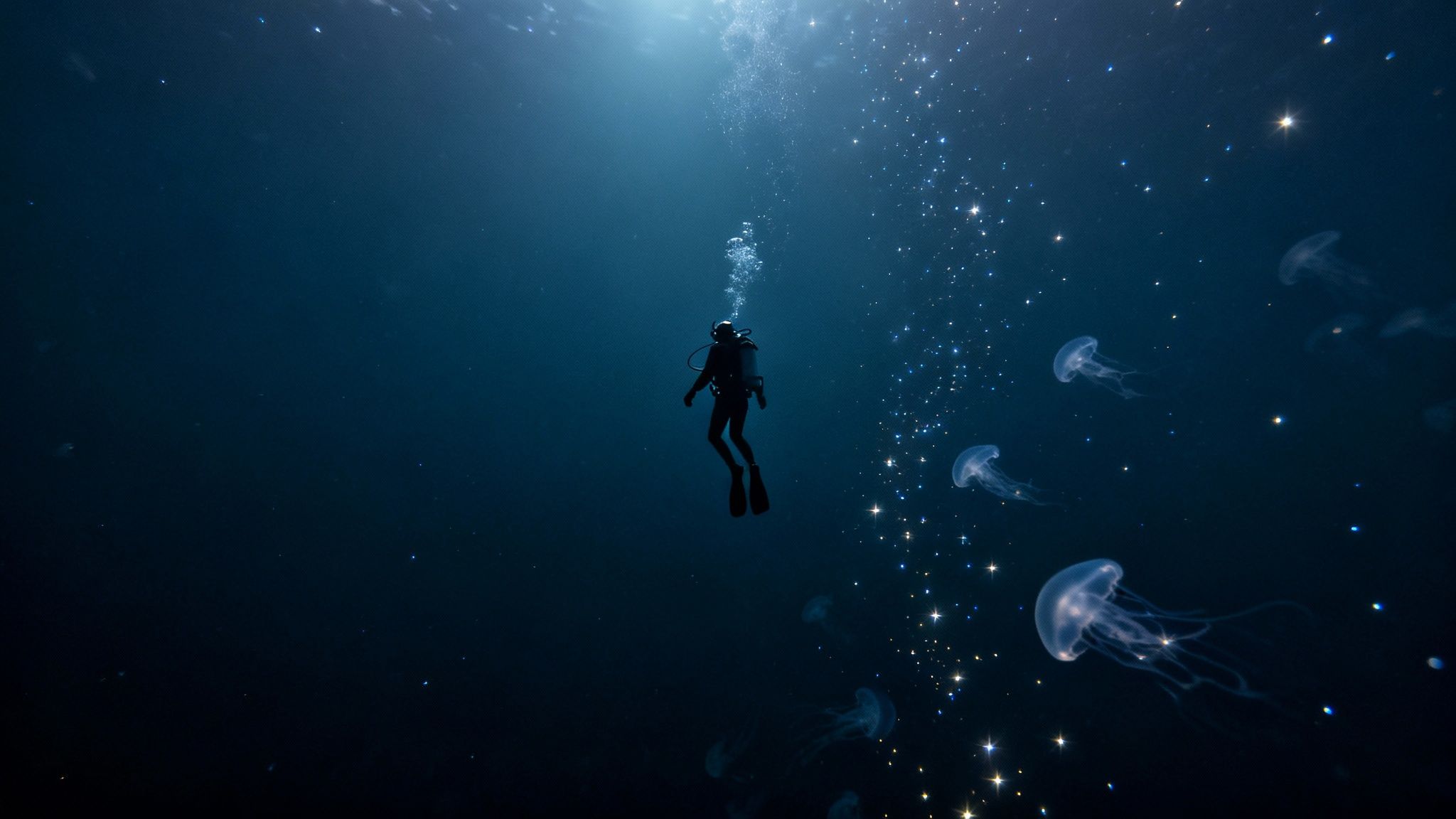 A diver floats in dark blue water surrounded by glowing jellyfish and shimmering light.