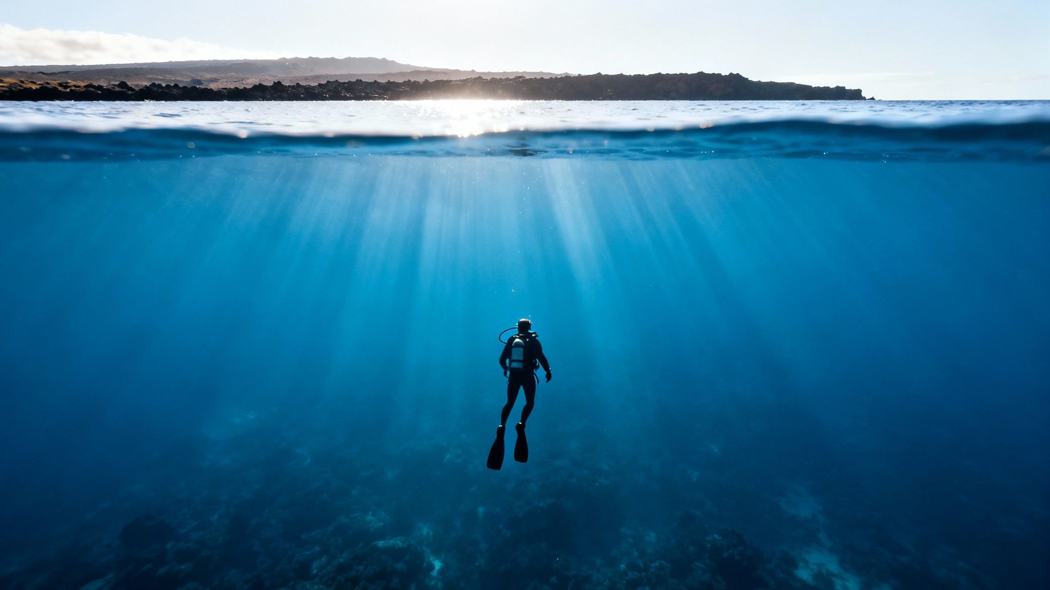 A scuba diver explores deep blue ocean waters with sun rays, and an island visible above the surface.