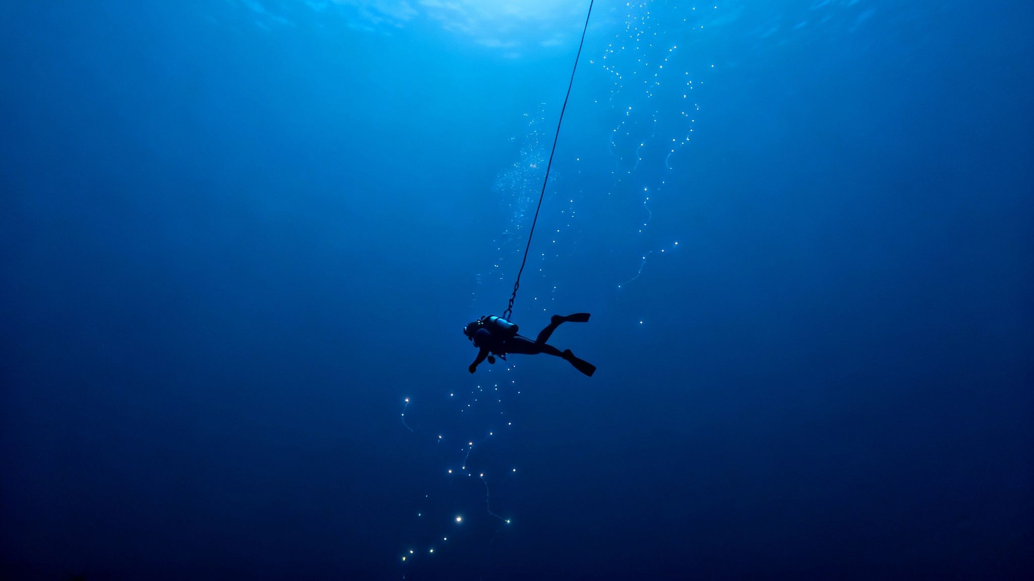 A lone scuba diver descends into the deep blue ocean, surrounded by light trails.