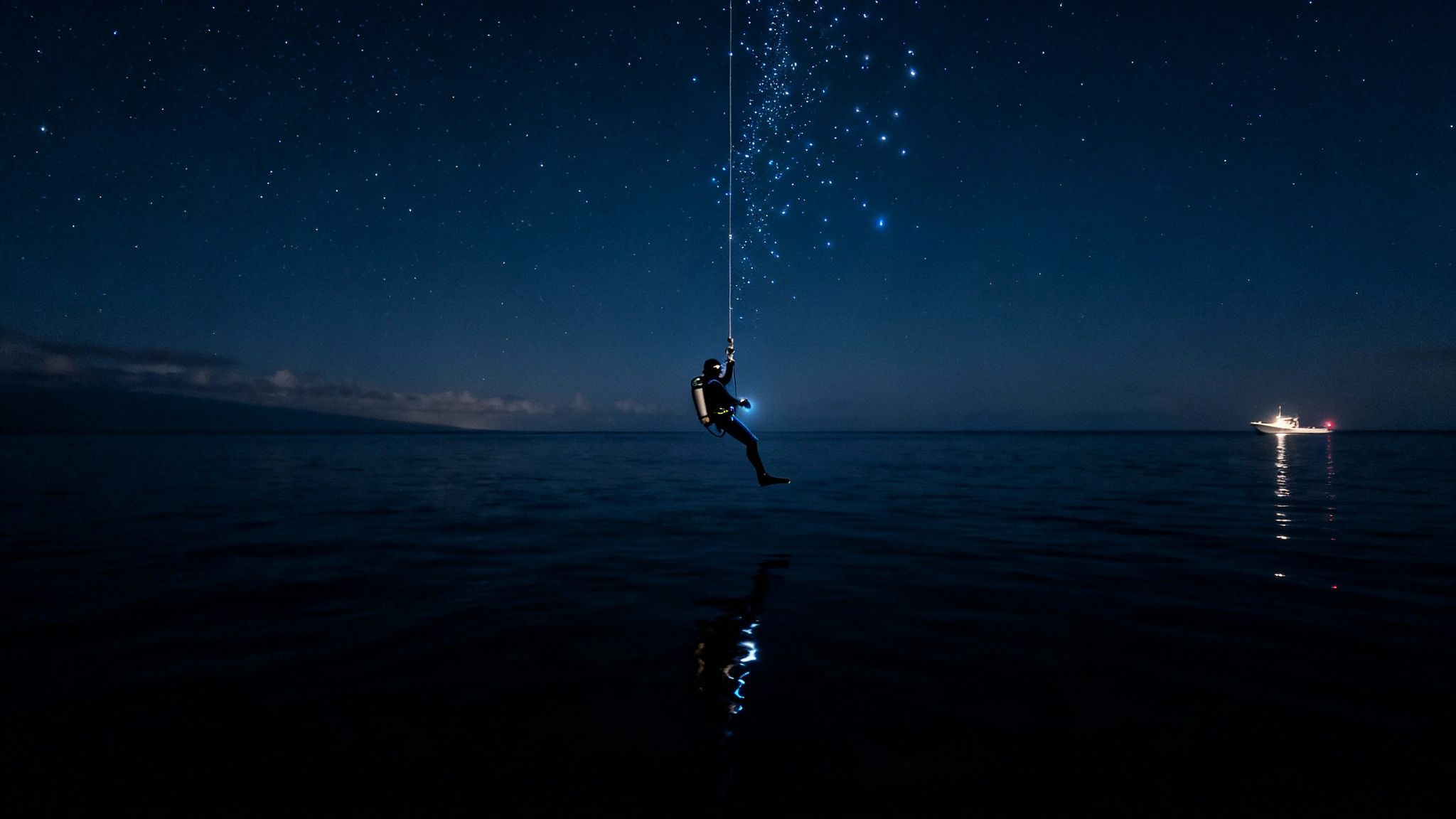 A diver suspended from a rope, illuminated by a glowing string of lights, over dark ocean water under a starry night sky. A boat with lights is visible in the distance.