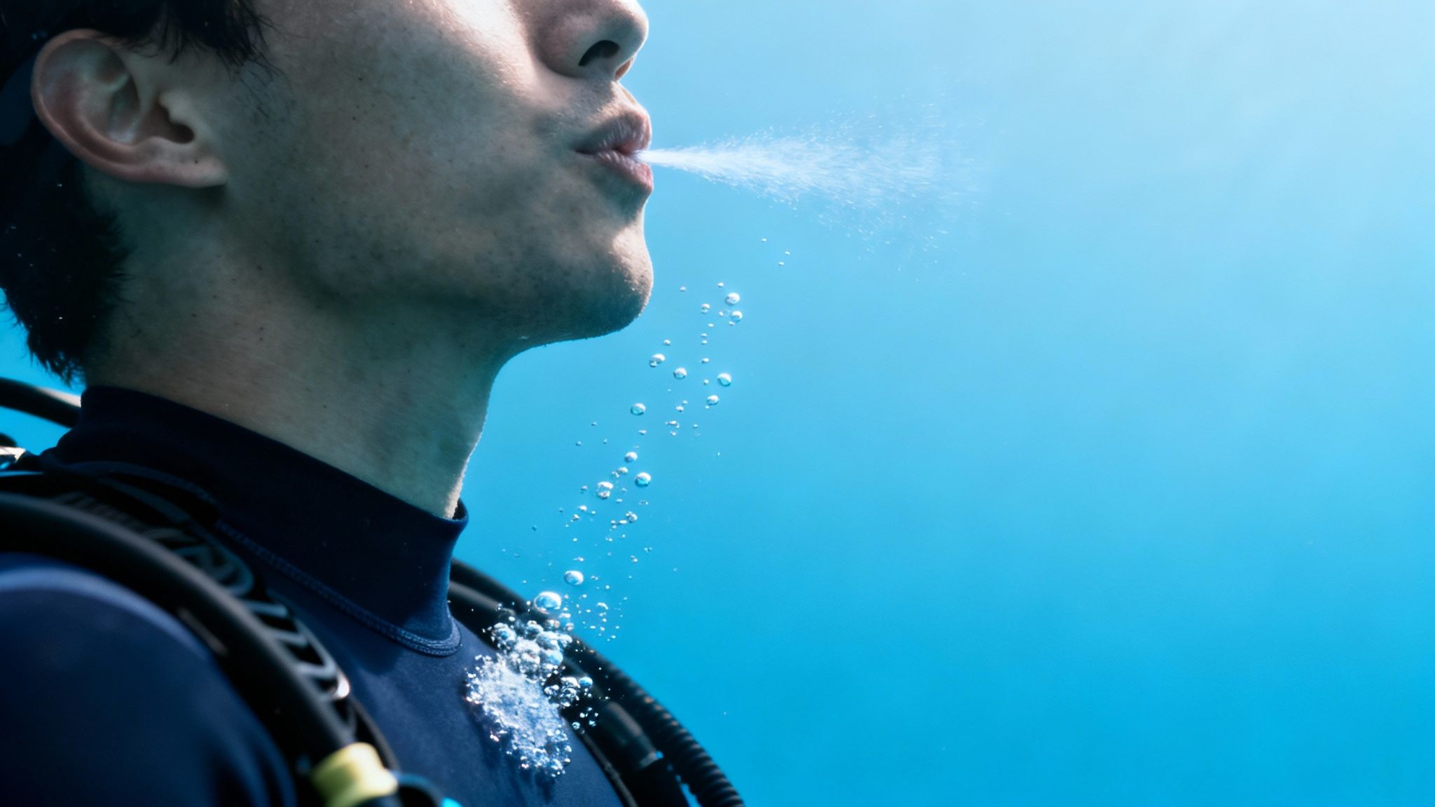 Close-up of a diver exhaling underwater, with bubbles rising against a bright blue background.