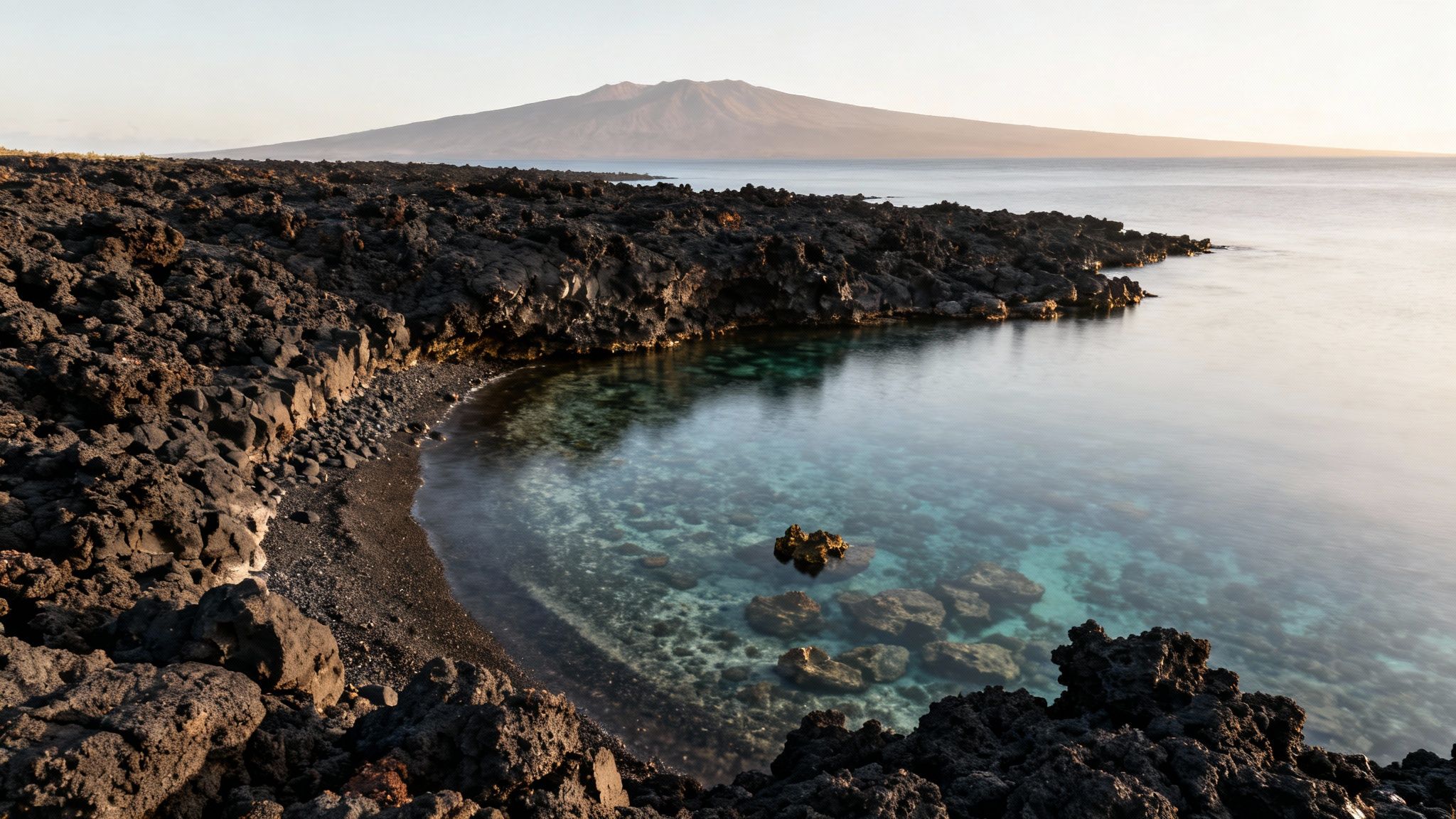 A serene volcanic coastline with clear turquoise water in a cove, overlooking a distant large island.