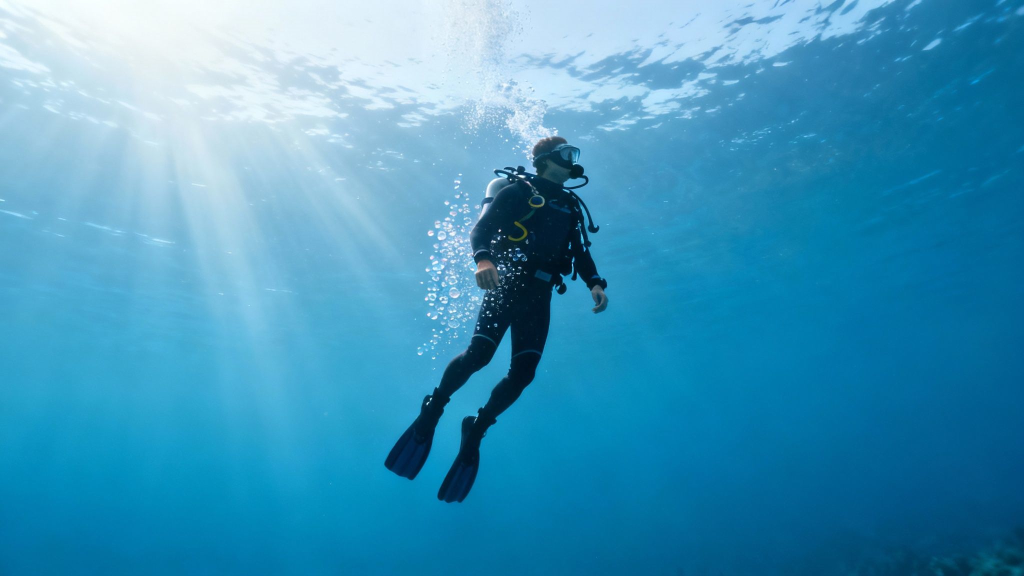 A scuba diver in a wetsuit ascends through clear blue water, performing a safety stop beneath sun rays.