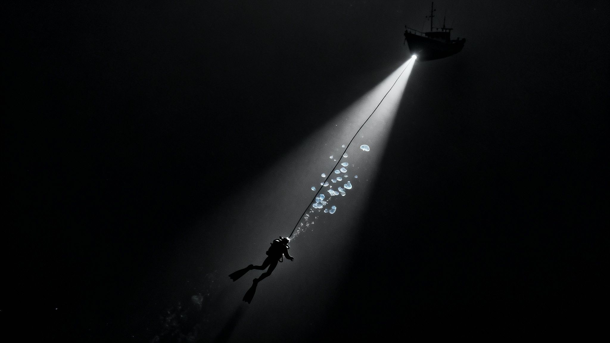 A diver descends into dark water, illuminated by a boat's spotlight, with air bubbles rising.