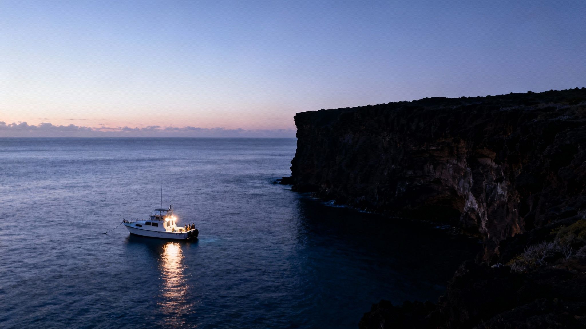 A brightly lit boat in dark, calm ocean water next to towering black cliffs at twilight.