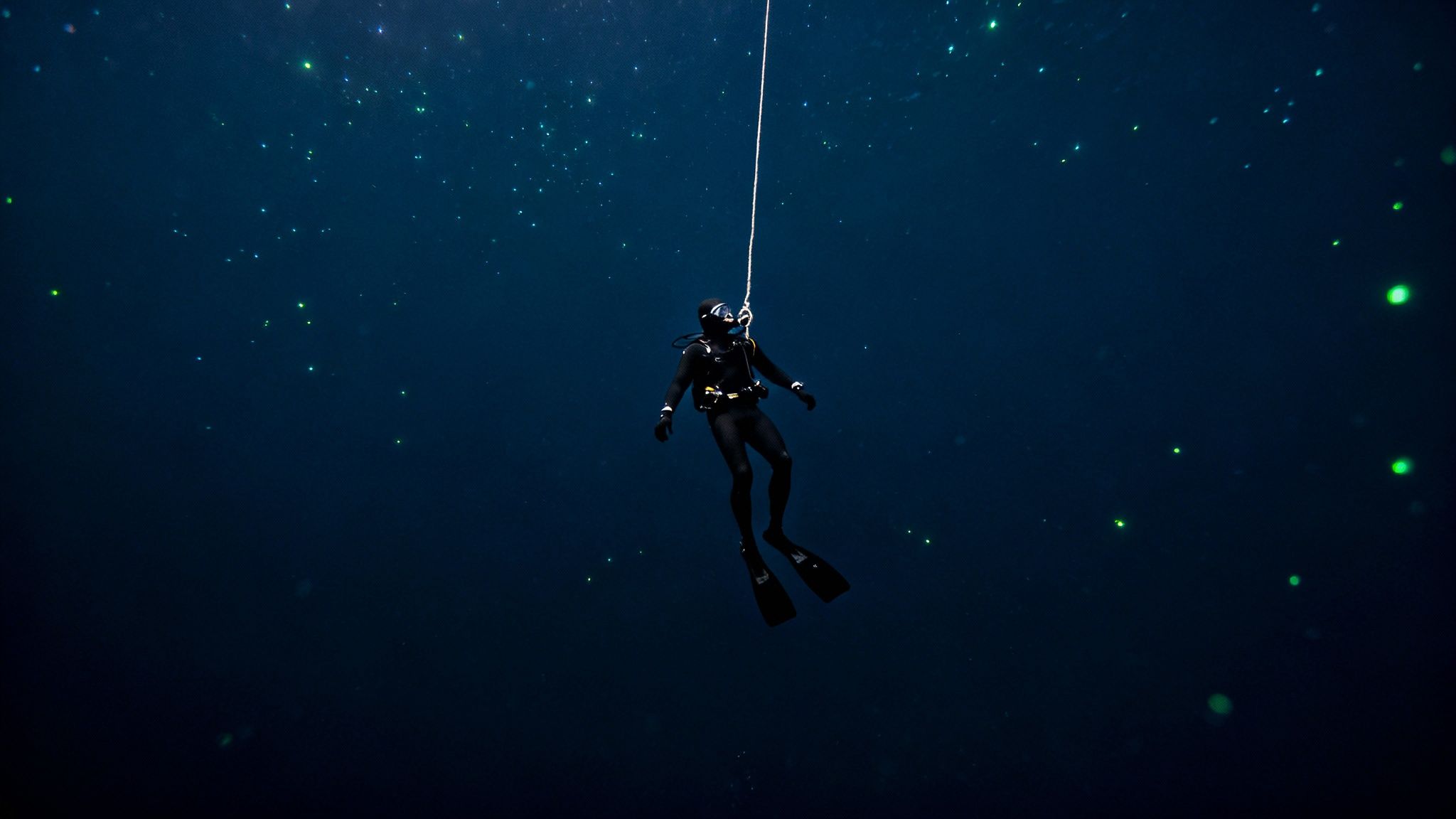 A free diver descends into deep blue water, tethered to a line, surrounded by glowing green bioluminescence.